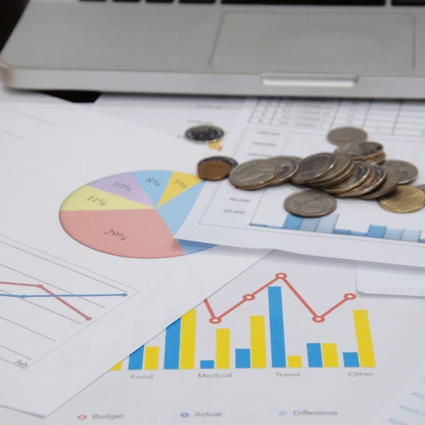 Coins are stacked on top of financial documents featuring colorful charts and graphs, with a laptop keyboard visible in the background—illustrating key data used in financial services. The papers show a pie chart, bar graphs, and a line graph.