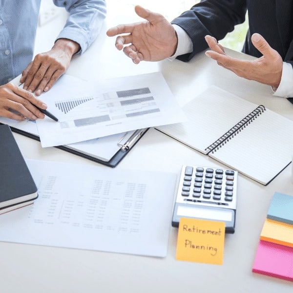 Two people discuss financial charts at a desk with a calculator, notebook, and documents. A sticky note labeled “Retirement Planning” suggests a meeting about future finances and exploring financial services options.