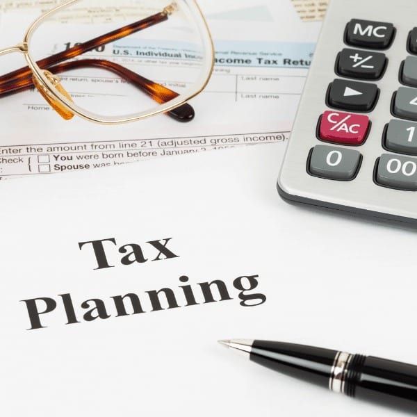 A pair of glasses, a calculator, and a pen rest on documents with the words "Tax Planning" prominently displayed, suggesting financial services or tax preparation.