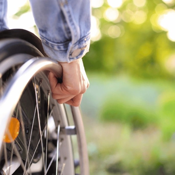 Close-up of a person’s hand on the wheel of a wheelchair, outdoors with greenery blurred behind. The individual, wearing a light blue denim jacket, considers insurance services from Spectre Financial for added peace of mind.