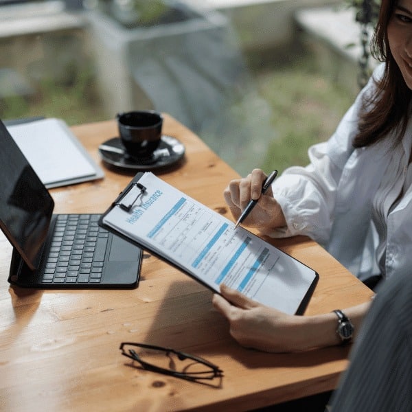 Two people sit at a wooden table with a laptop, notebook, and coffee. One holds a clipboard with a health insurance form from Spectre Financial, while the other writes on it—discussing insurance services over coffee.