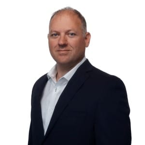 A man with short light hair wearing a dark suit jacket and a white dress shirt stands against a plain white background, looking directly at the camera with a neutral expression, representing financial services Windsor.