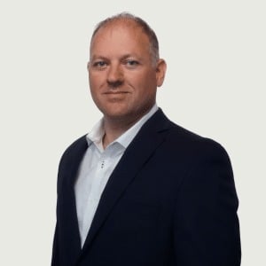 A man with short light hair, dressed in a dark suit jacket over a white collared shirt, stands facing slightly to the side against a plain, light background—conveying the professionalism often seen in financial services.