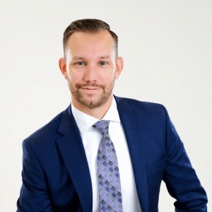 A man with short, neatly styled hair and a trimmed beard is wearing a blue suit, white shirt, and patterned tie. He is smiling slightly and facing the camera against a plain, light background—ideal for financial services Windsor professionals.
