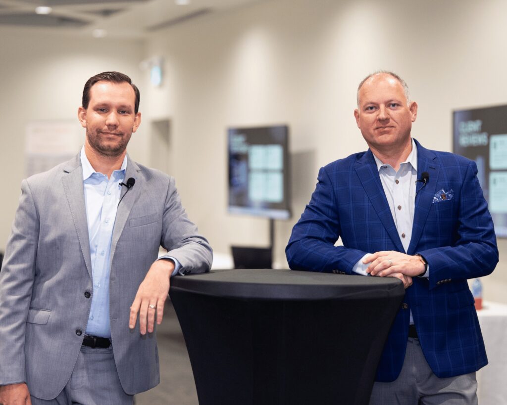 Two men in business suits stand beside a tall round table in a modern conference room, both wearing clip-on microphones, discussing Financial Events & Community Initiatives by Spectre Financial. Blurred screens and lights are visible in the background.