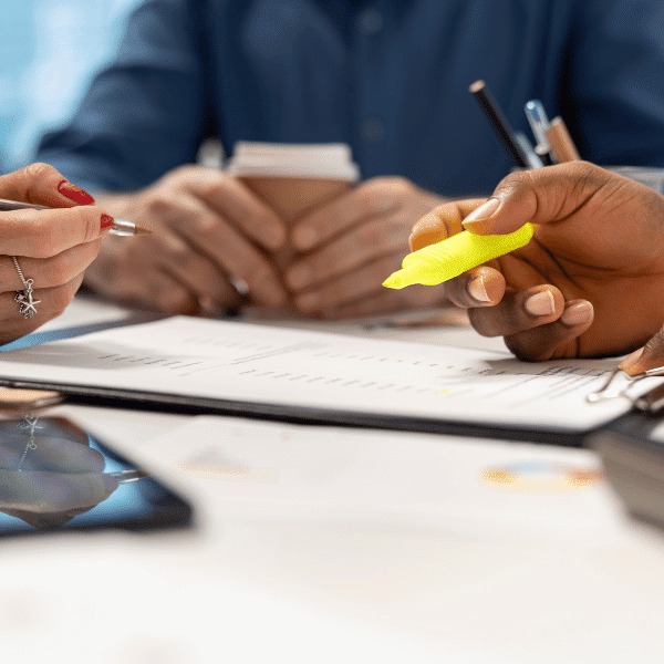 Close-up of three people at a desk, including an insurance broker, holding pens and a highlighter, working together on documents with coffee cups and a tablet visible, suggesting a collaborative meeting or discussion.