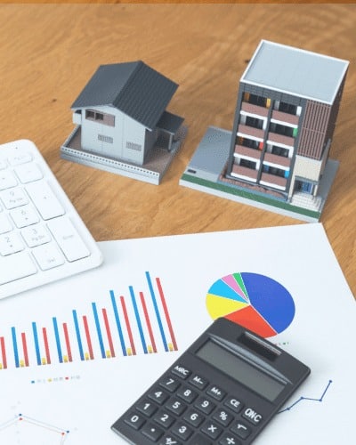 Two small model buildings, a keyboard, calculator, and financial charts with graphs and a pie chart are arranged on a wooden desk, highlighting real estate analysis or investment planning tied to financial services in Windsor.