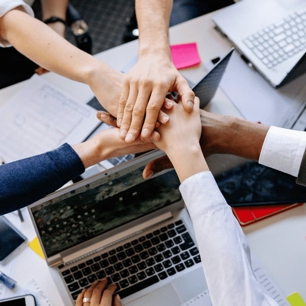 Several people, including an insurance broker, stack their hands together in a gesture of teamwork over a table covered with laptops, papers, and office supplies. The image conveys collaboration and unity in a work environment.