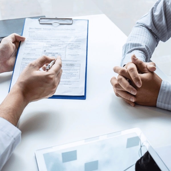 Two people sit at a table; an insurance broker holds a clipboard with a form and pen, gesturing as they explain, while the other person sits with hands clasped. A tablet and paper are also visible on the table.