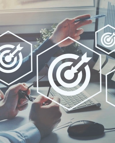 Two people sit at a desk with a keyboard, mouse, and glasses, discussing work. Overlay graphics of target icons with arrows symbolize goals, focus, and business strategy in the financial services Windsor sector.
