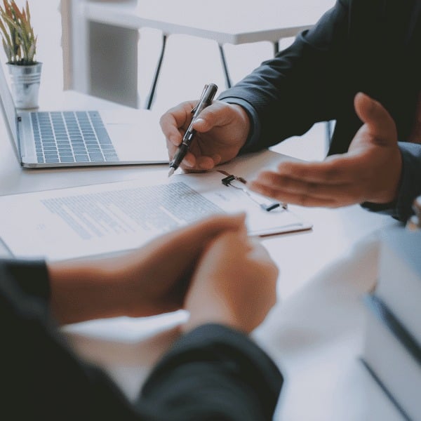 Two people sit at a desk with a laptop, documents, and a pen. One person is signing a paper while an insurance broker looks on, suggesting a formal business meeting or contract signing.